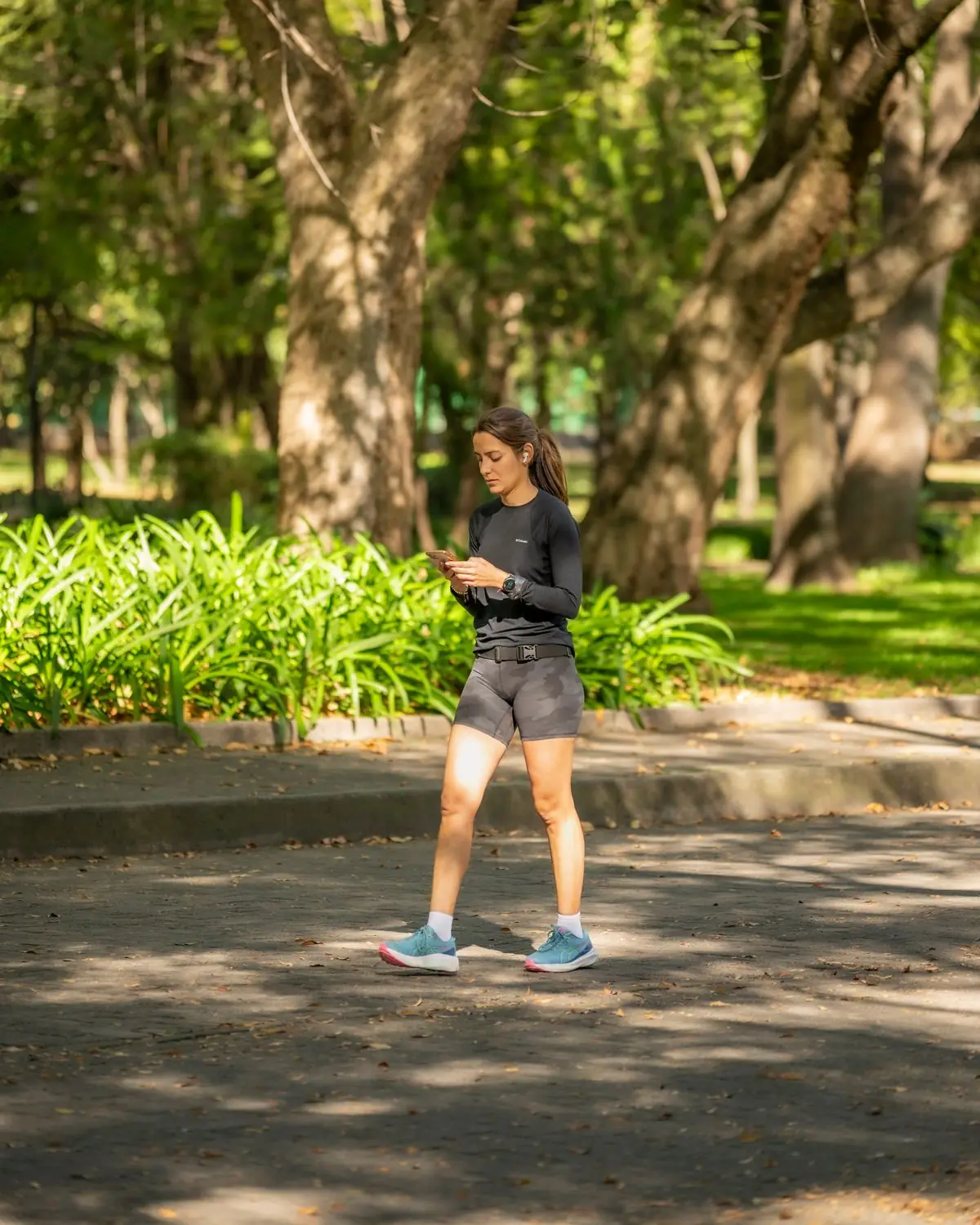 Woman jogging on pavement in a sunlit park surrounded by trees and greenery.