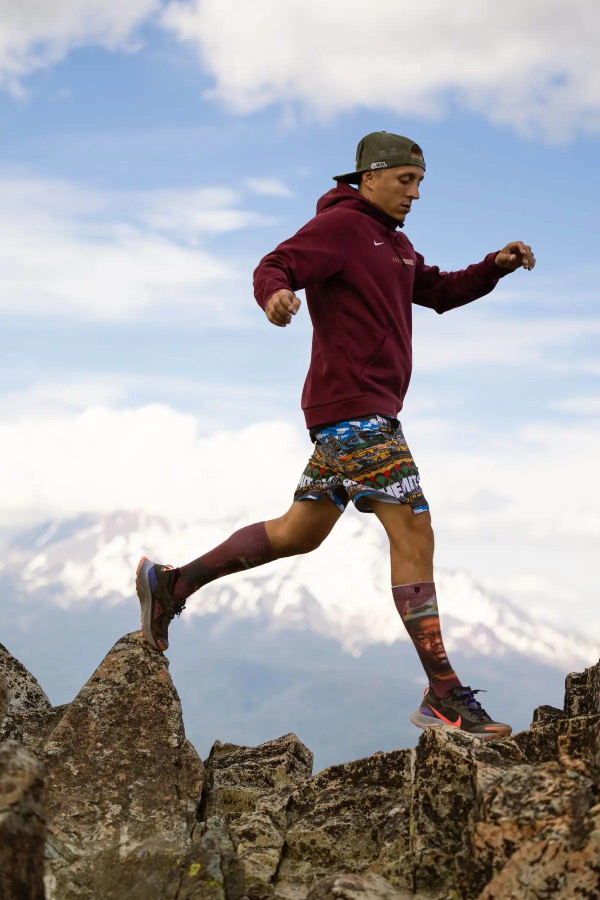 A young man in athletic gear is seen trail running over rugged mountain terrain with a serene backdrop.
