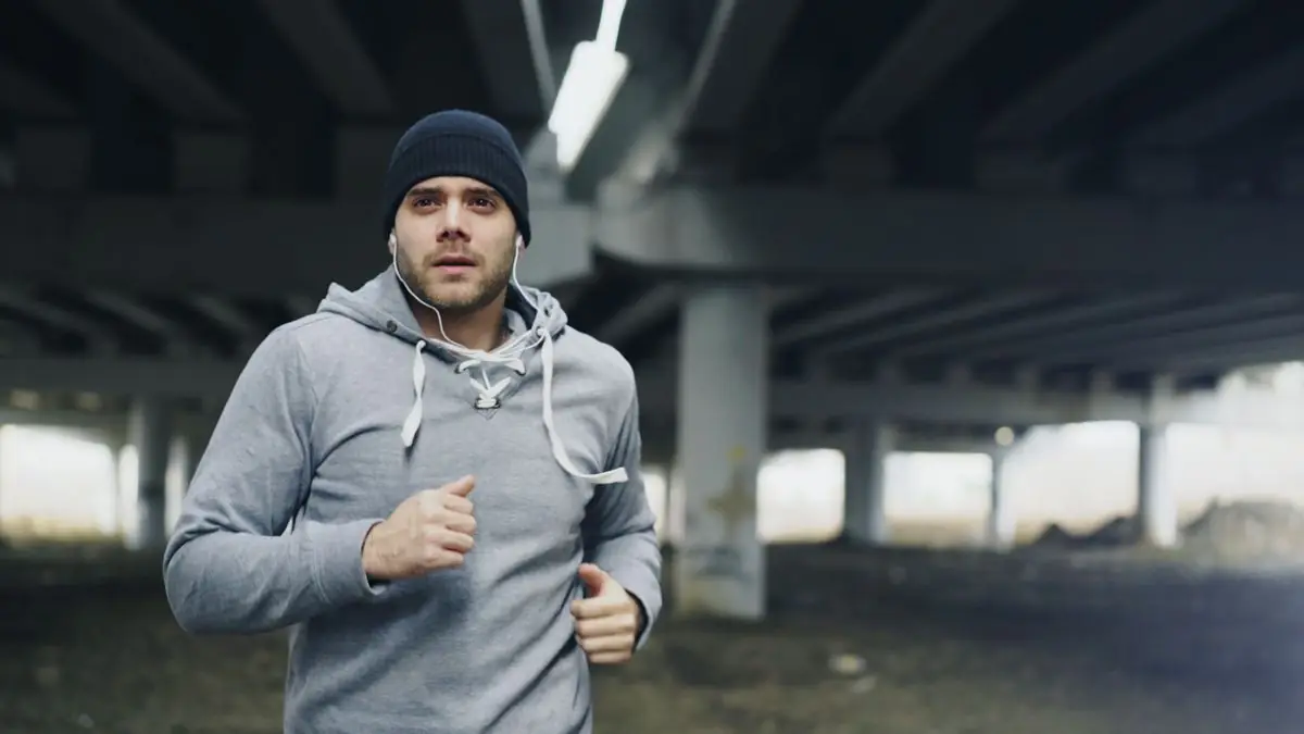 A man in athletic wear jogging under a city bridge.