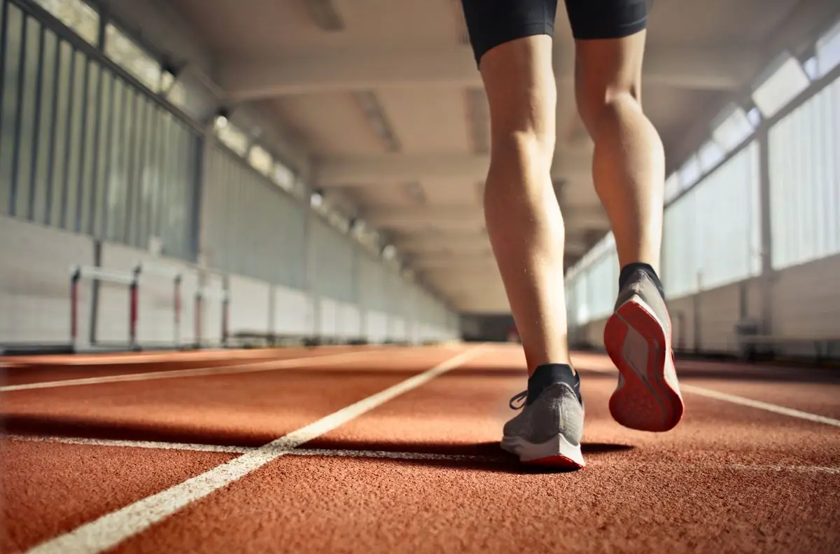 Back view of crop strong runner walking along running track in athletics arena while doing warm up exercises during workout.