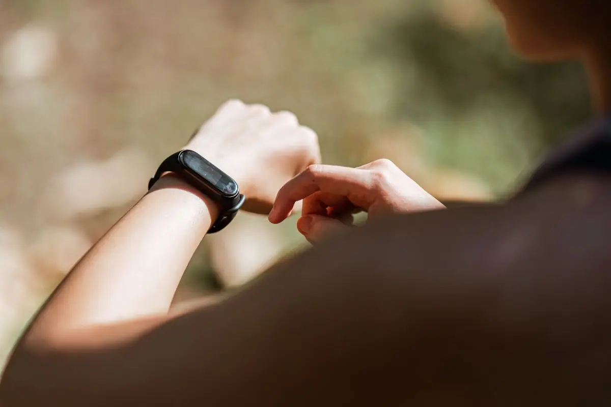 Crop anonymous sportswoman checking notifications on modern fitness tracker while training against blurred nature background on sunny weather.