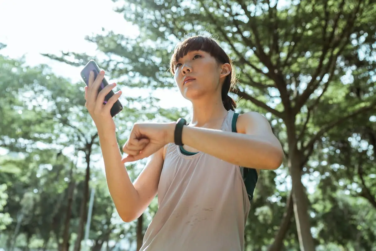 Female wearing sport clothes standing in park and connecting smartphone and smart watch during break in sunny day.