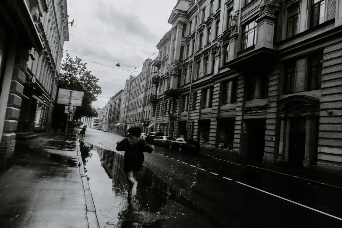 Black and white full body of anonymous person running on puddles on road along residential buildings.