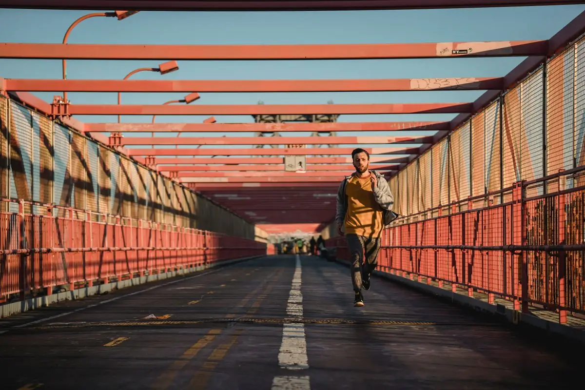Energetic male running on asphalt path on pedestrian footbridge under metal structure during training on sunny street.