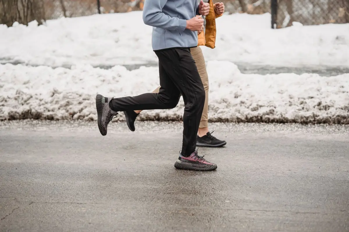 Side view crop anonymous joggers in sportswear running together on road in snowy park on cold winter weather.
