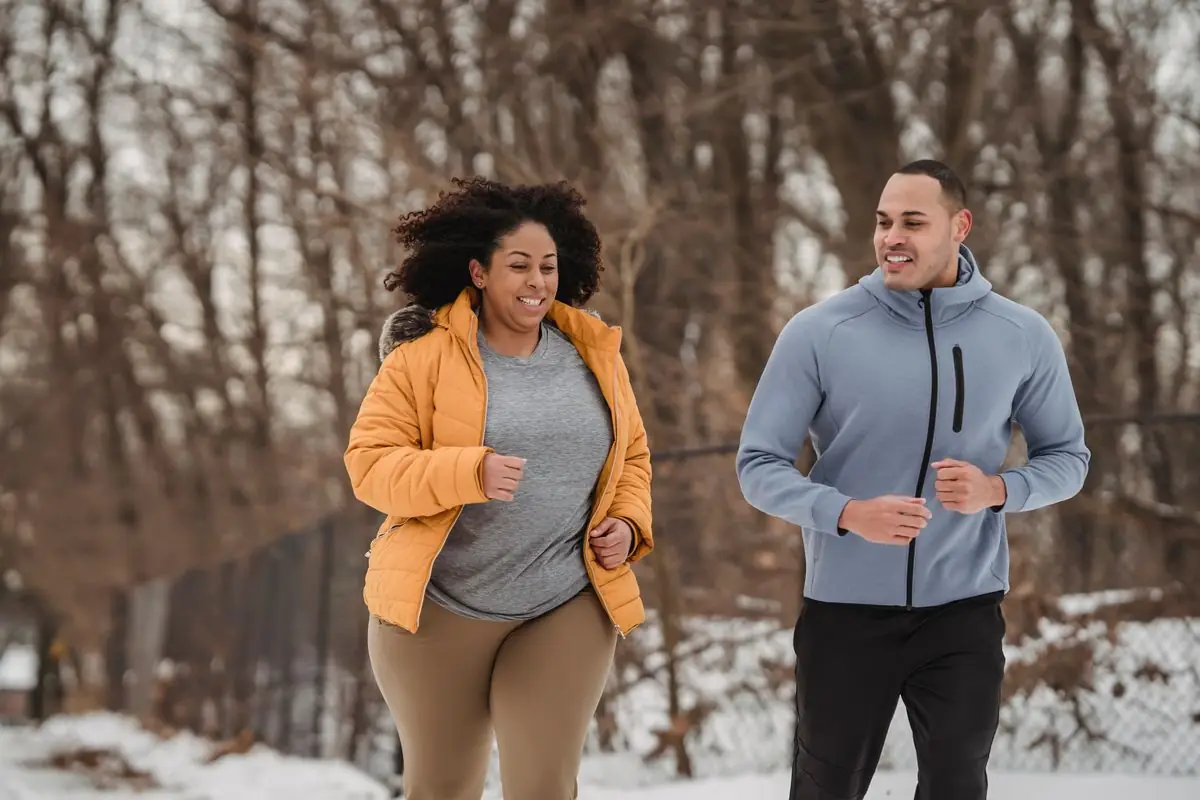 Happy fit male trainer in activewear jogging with smiling black female in outerwear in cold winter park.